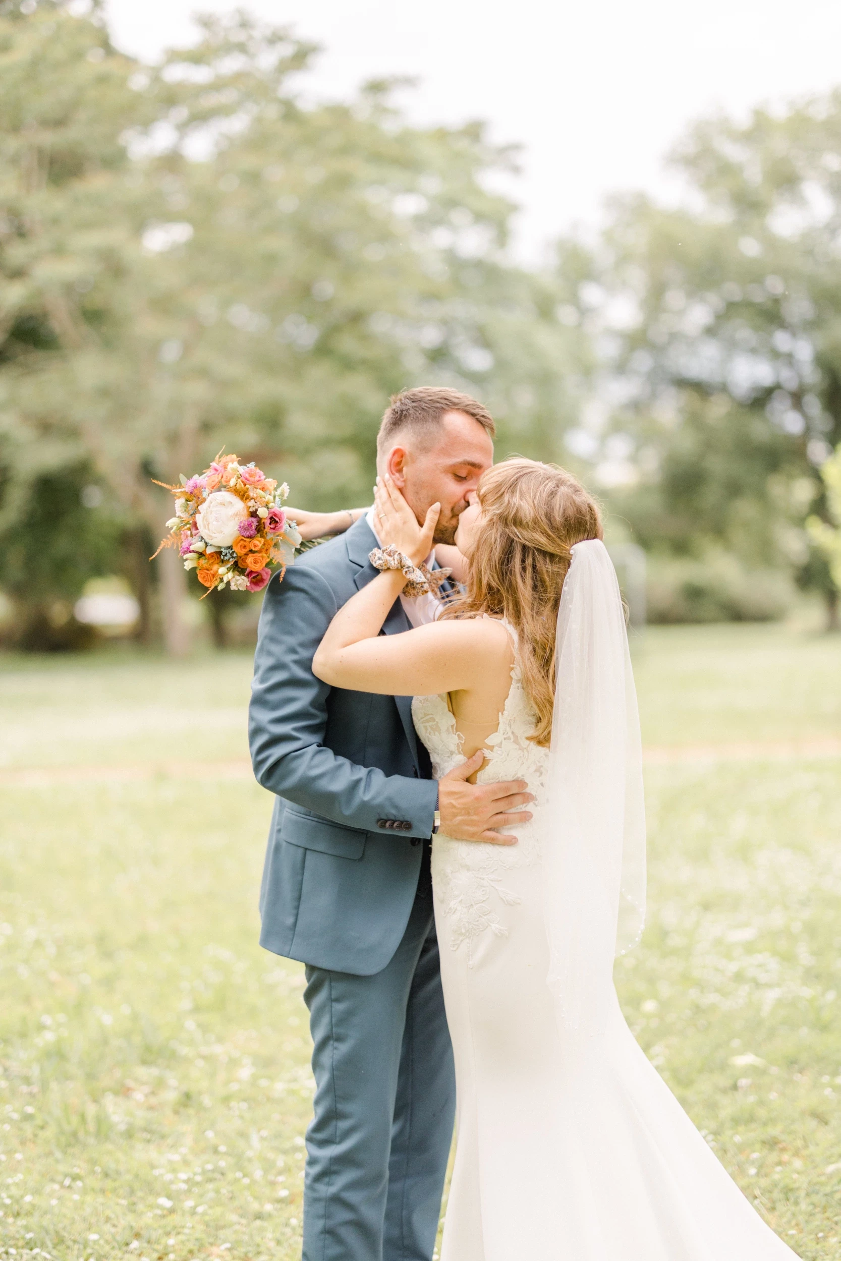 Couple de mariés s'embrassant tendrement dans un parc verdoyant, bouquet orange à la main