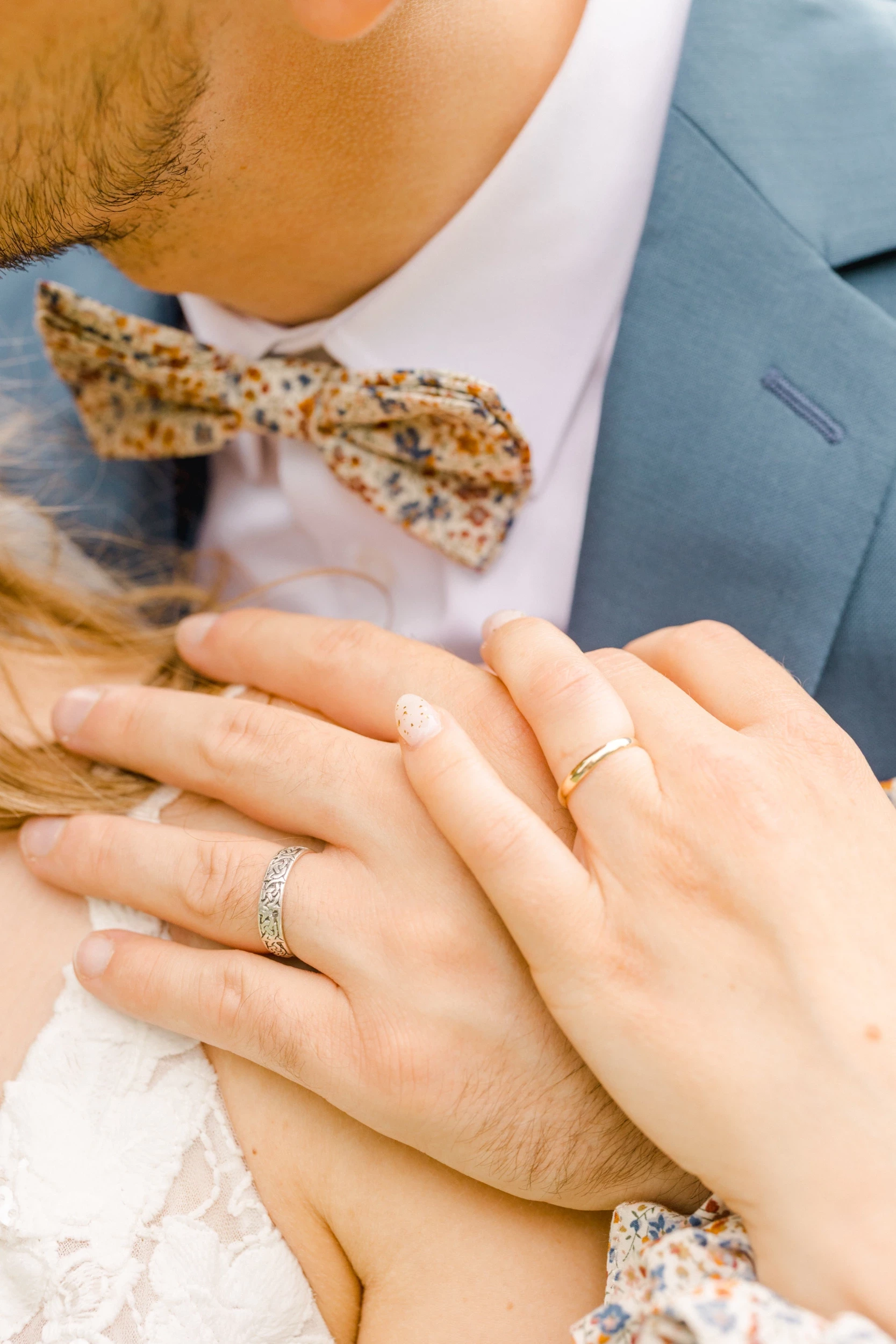 Gros plan sur les mains enlacées des mariés portant leurs alliances, noeud papillon fleuri visible