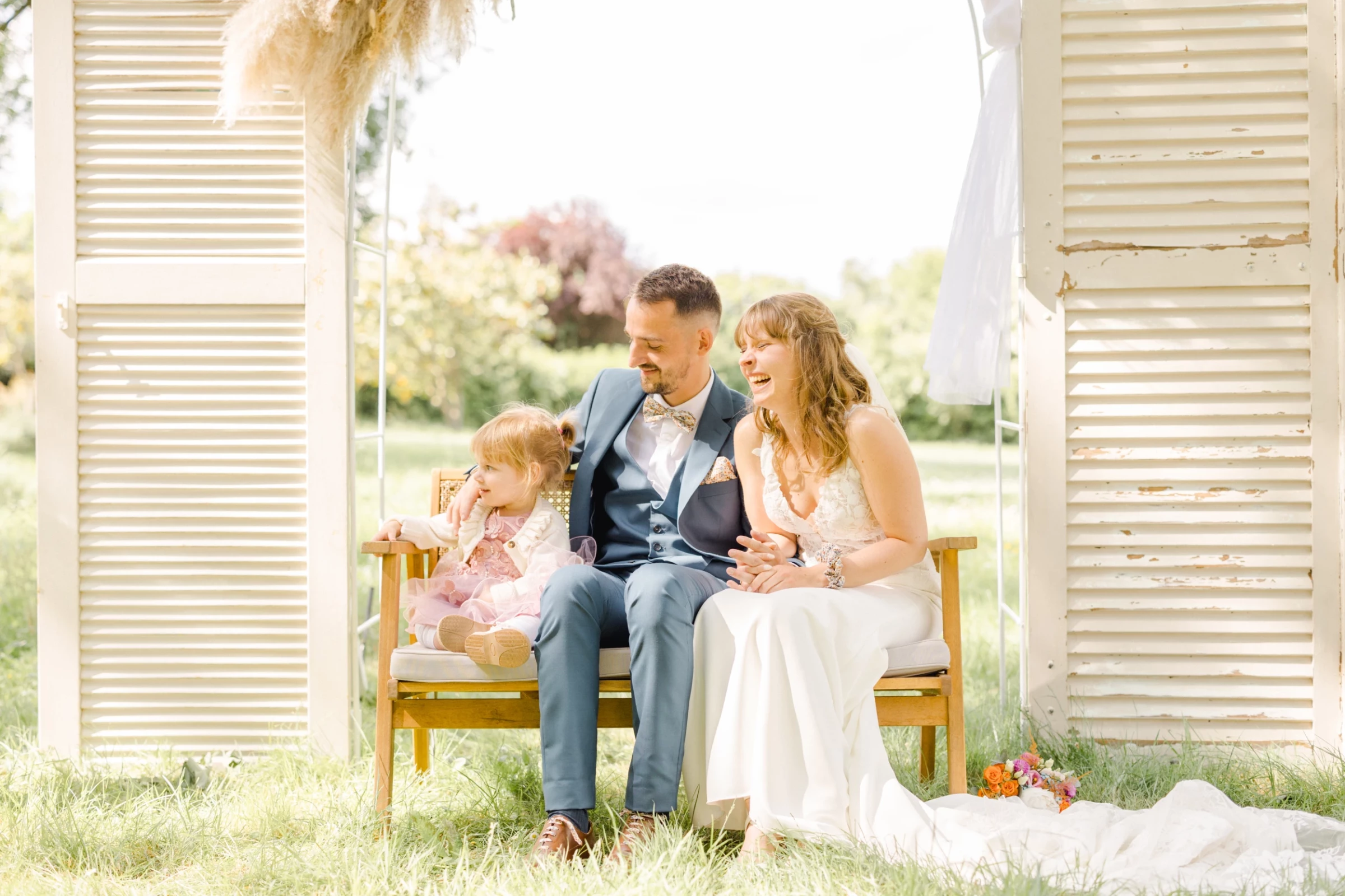 Jeune famille assise sur un banc vintage entre portes anciennes blanches dans un jardin champêtre