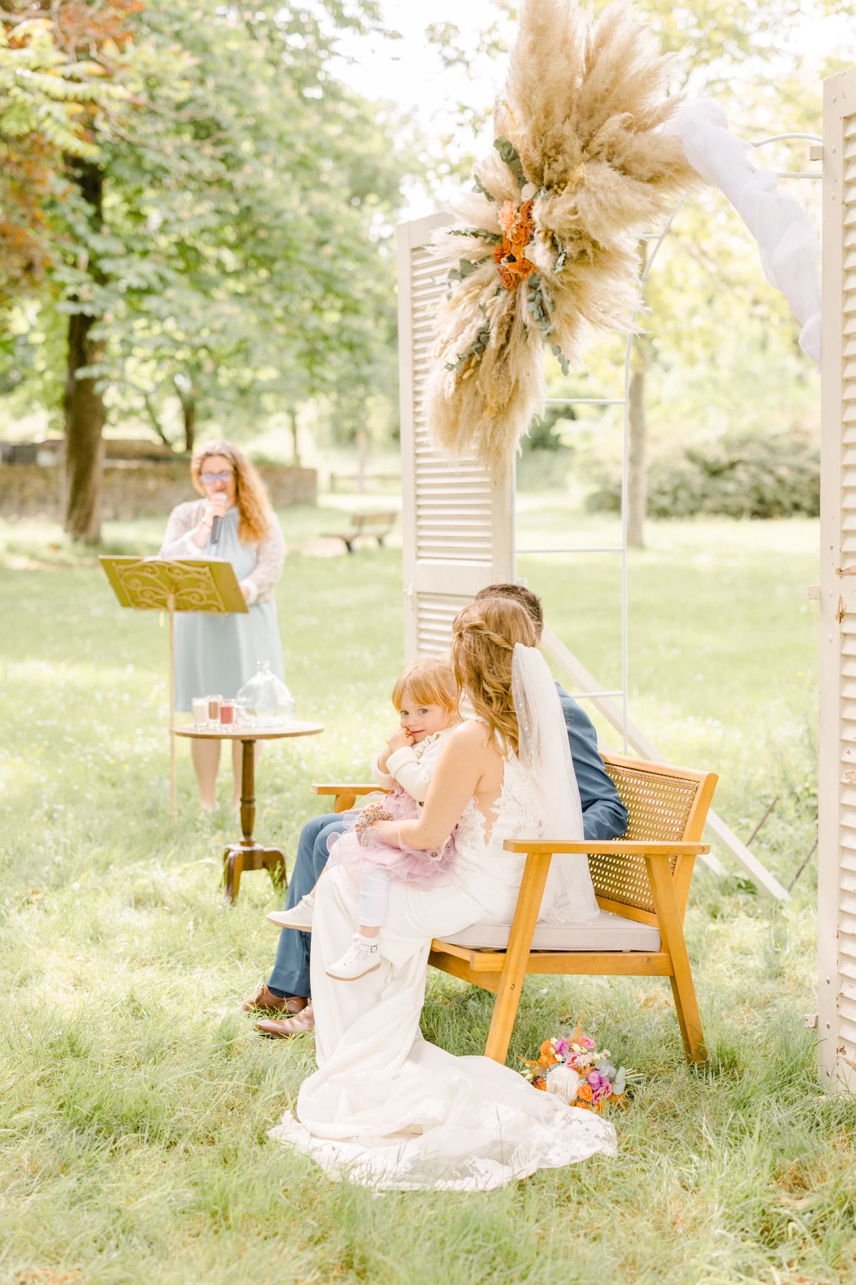Cérémonie laïque en plein air avec couple marié assis devant une arche fleurie et officiant lisant un texte