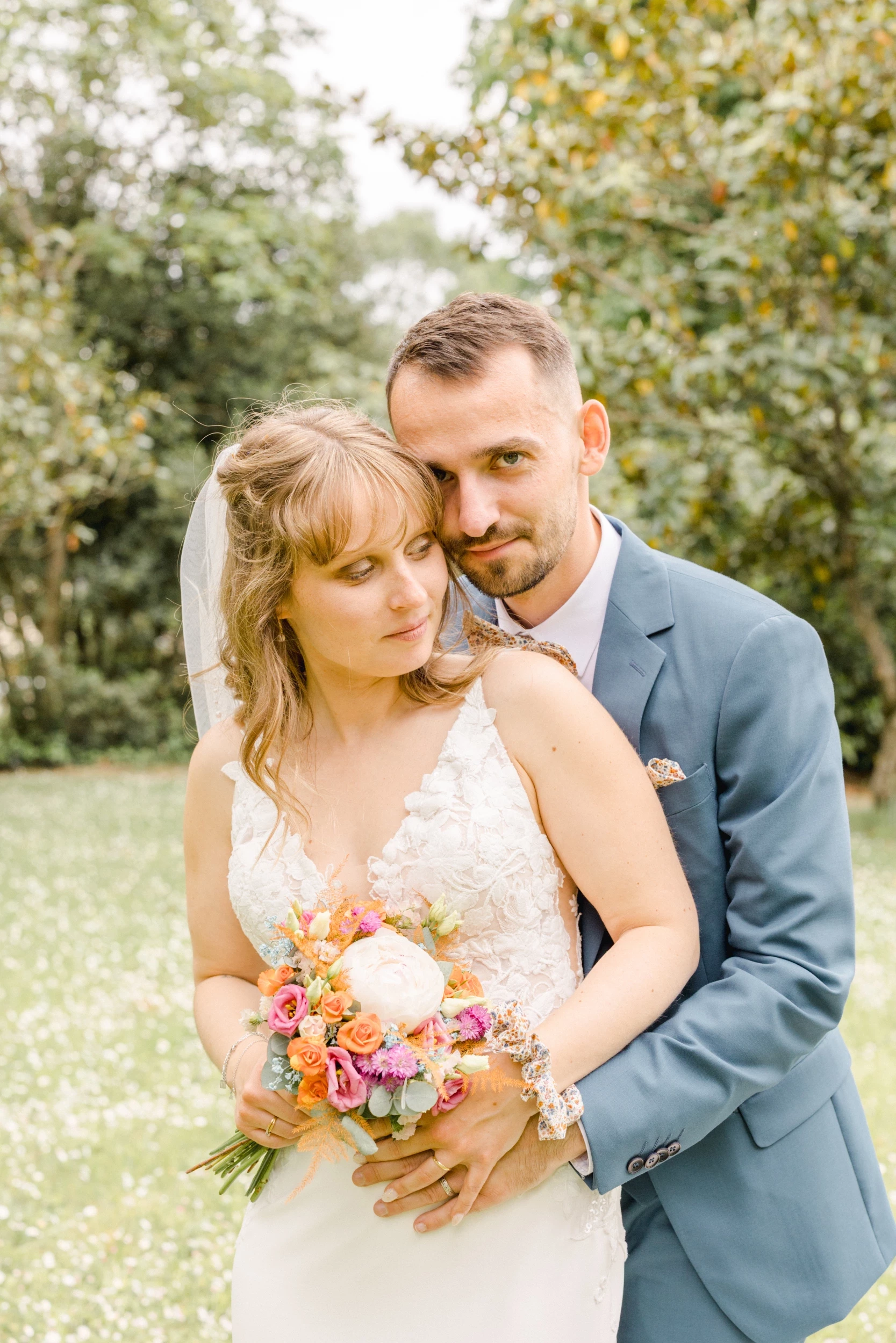 Couple de mariés enlacés dans un jardin verdoyant, la mariée tenant un bouquet de fleurs pastel