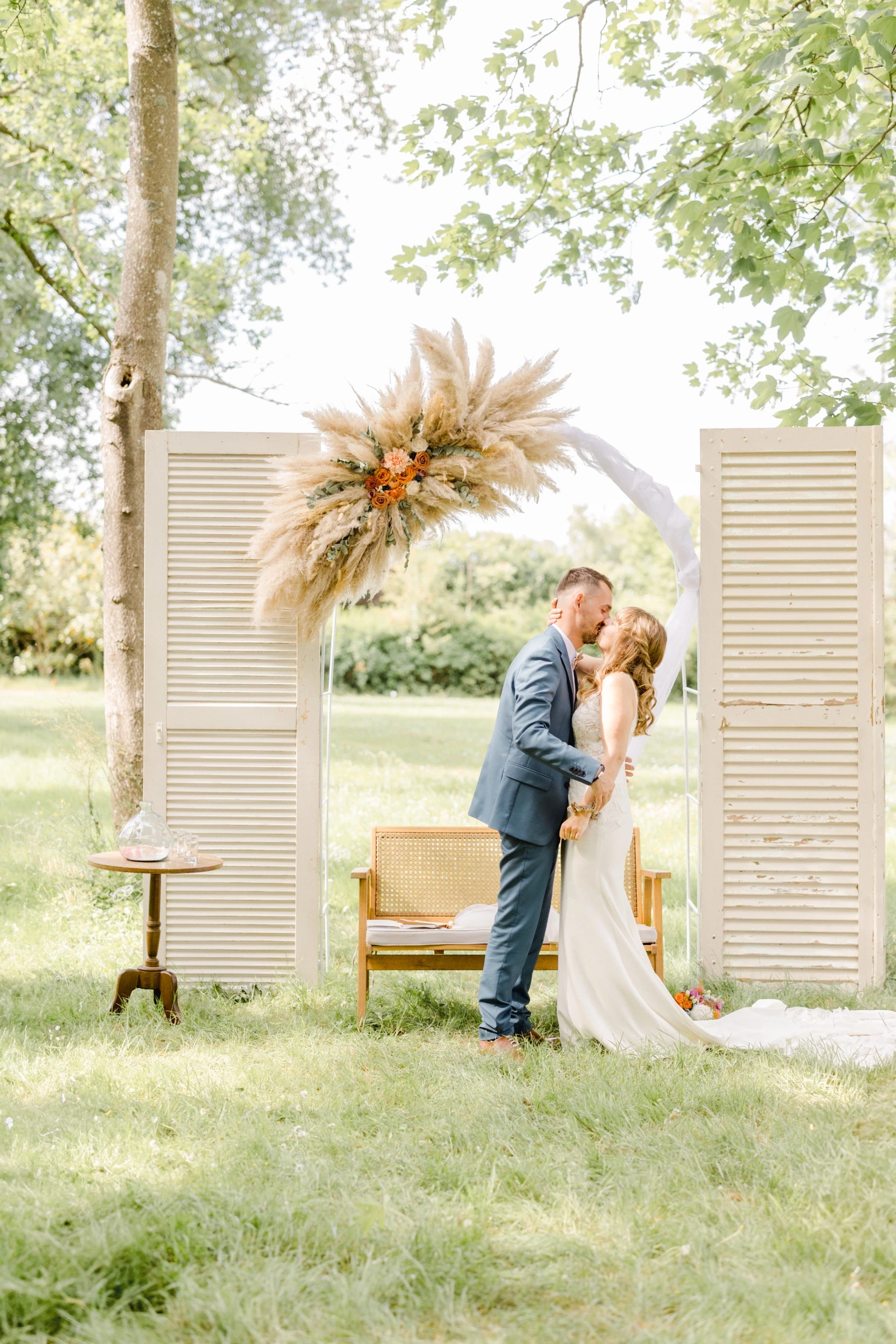 Couple de mariés s'embrassant devant des volets blancs et une arche de fleurs séchées en extérieur
