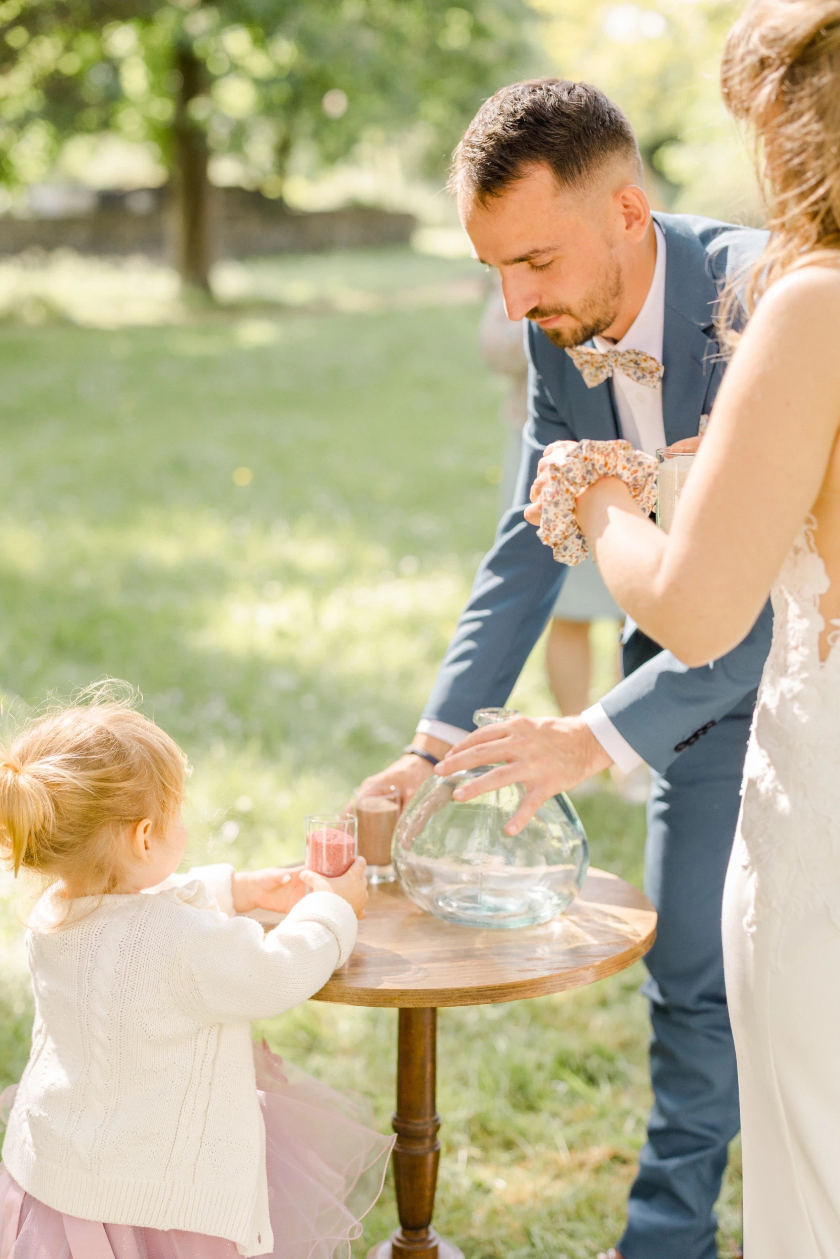 Couple de mariés et enfant versant du sable coloré dans un vase lors d'une cérémonie laïque en extérieur