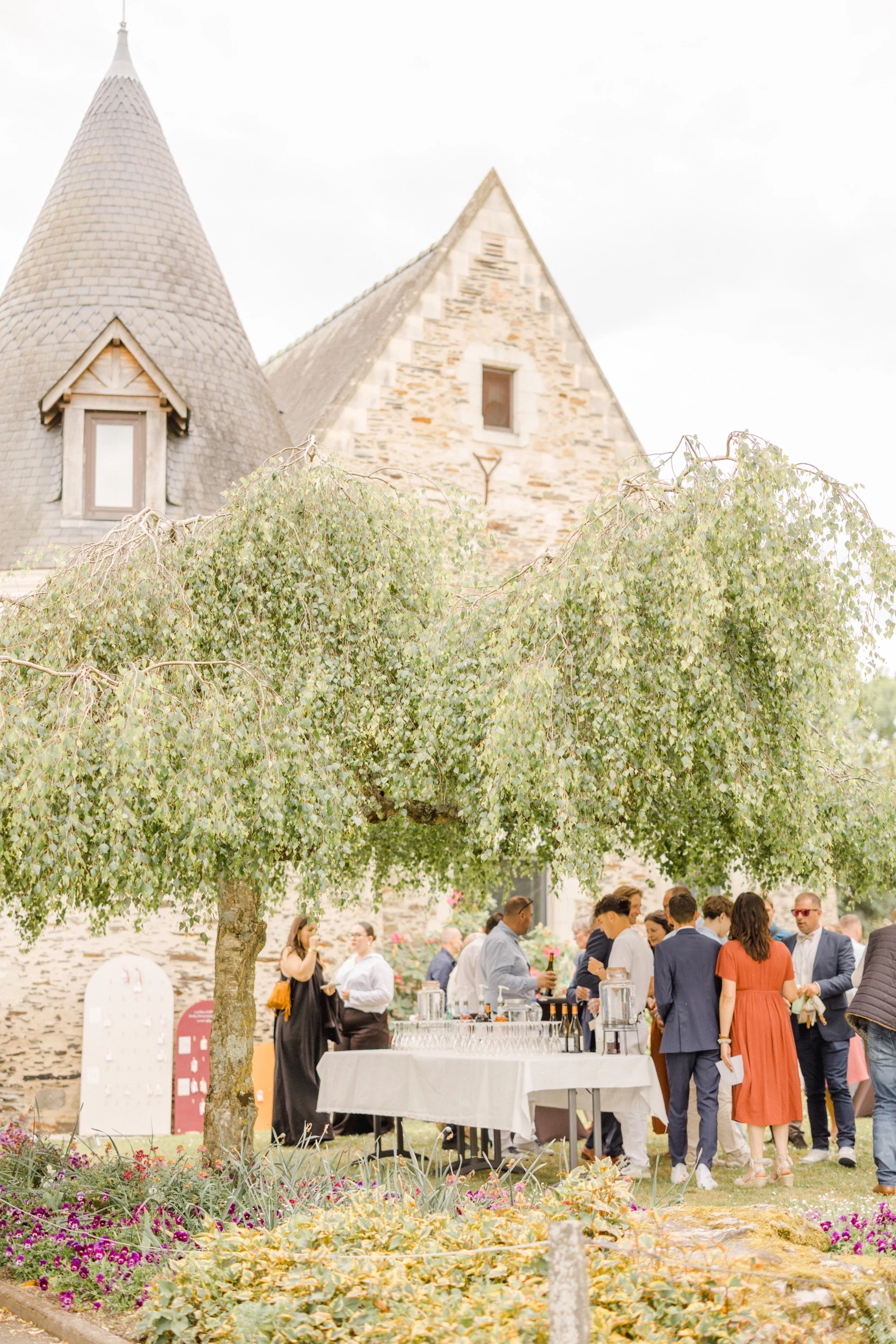 Cocktail de mariage sous un olivier devant un château en pierre aux toits coniques et murs beiges