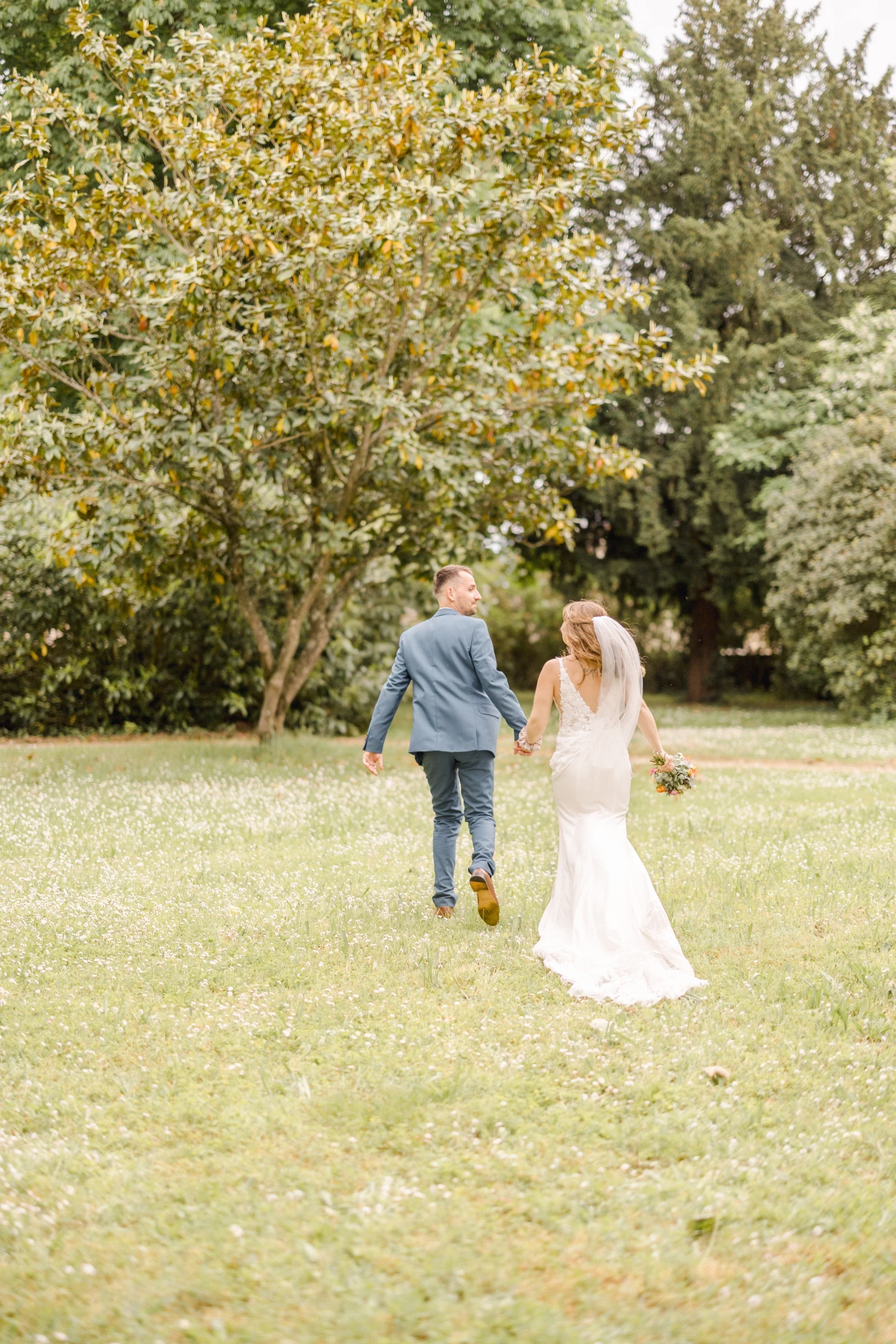 Couple de mariés courant main dans la main dans un pré verdoyant sous des arbres en fleurs