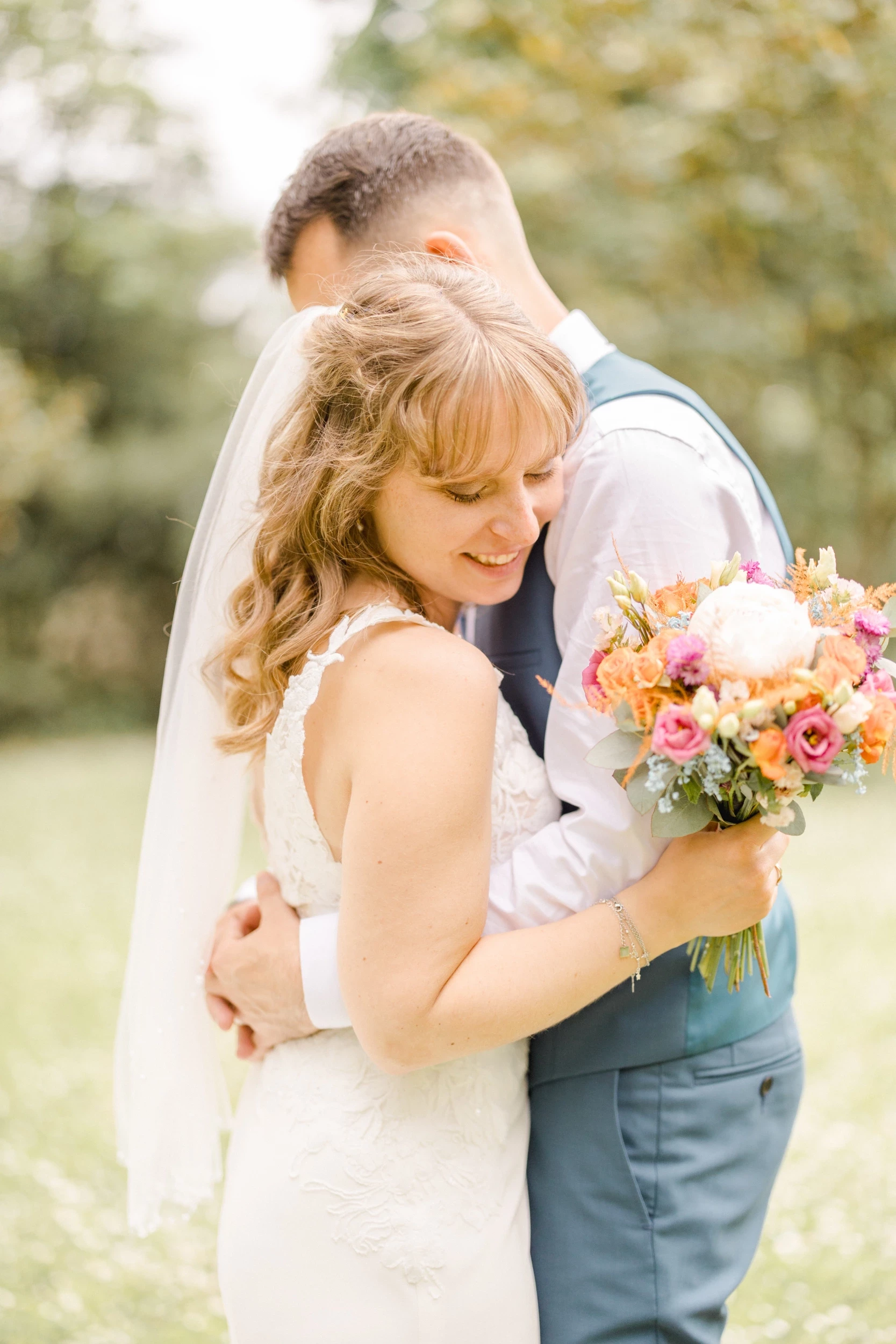 Mariée souriante enlacée par son époux, tenant un bouquet coloré dans un jardin ensoleillé