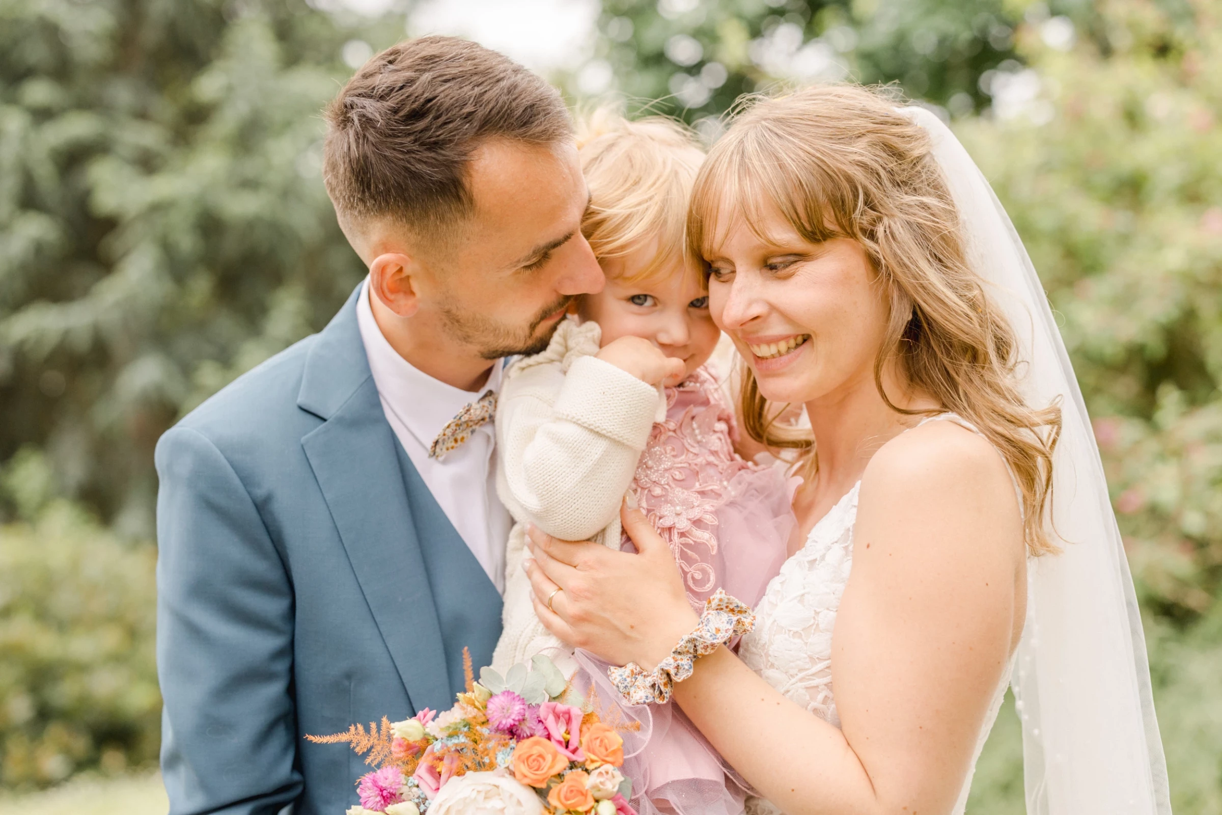 Couple de mariés enlace leur petite fille en robe rose lors d'un mariage en extérieur lumineux