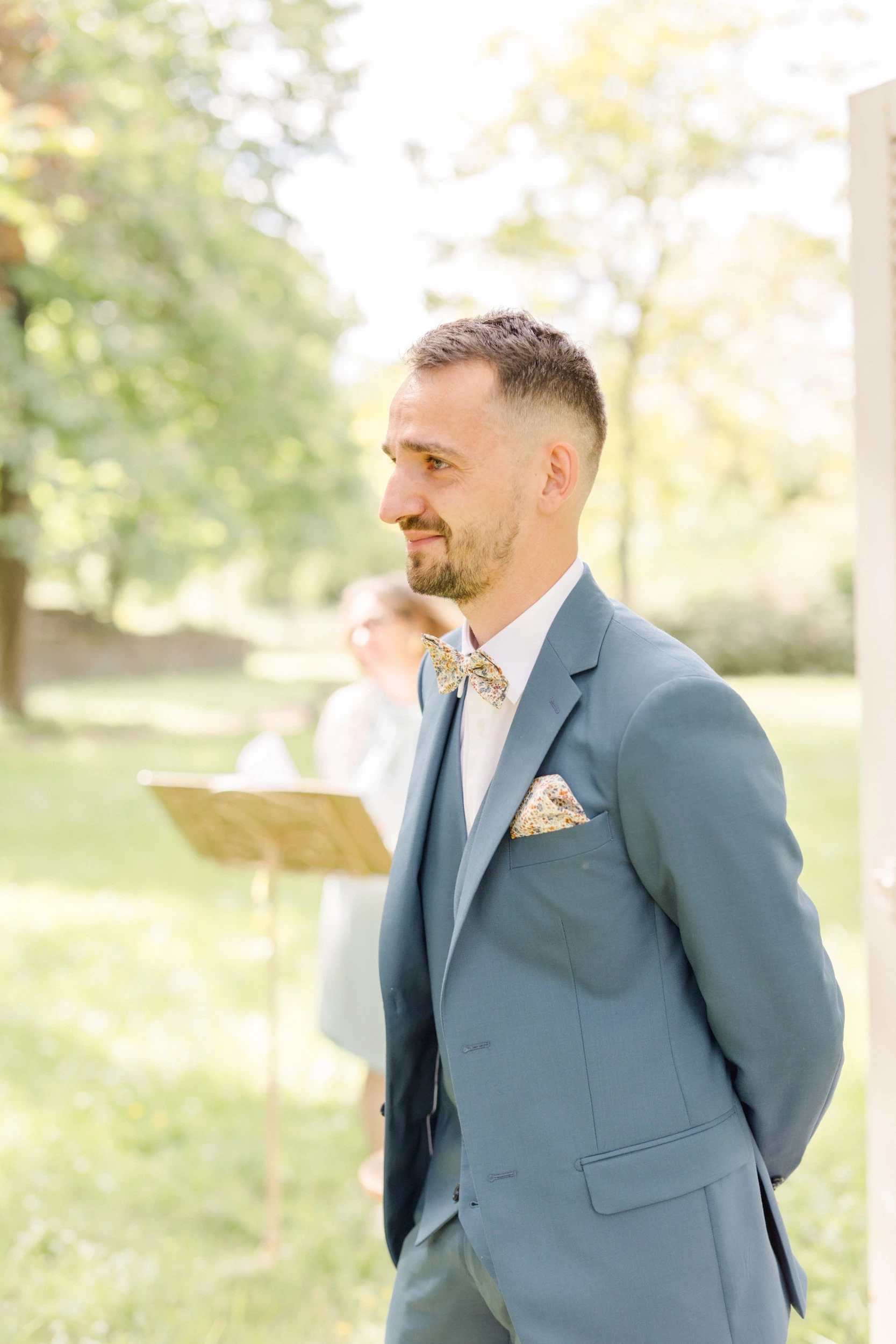 Marié en costume gris-bleu avec nœud papillon et pochette, souriant lors de sa cérémonie de mariage en extérieur