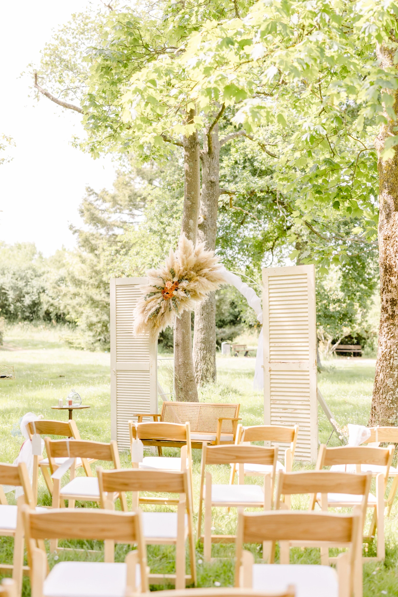 Cérémonie laïque en extérieur avec rangées de chaises en bois, arche fleurie dorée et portes blanches anciennes