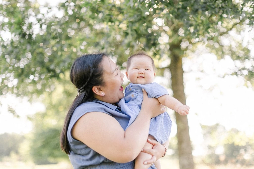 Séance photo famille en extérieur : mère tenant son bébé souriant dans ses bras, sous la lumière dorée des arbres, moment de tendresse et complicité authentique