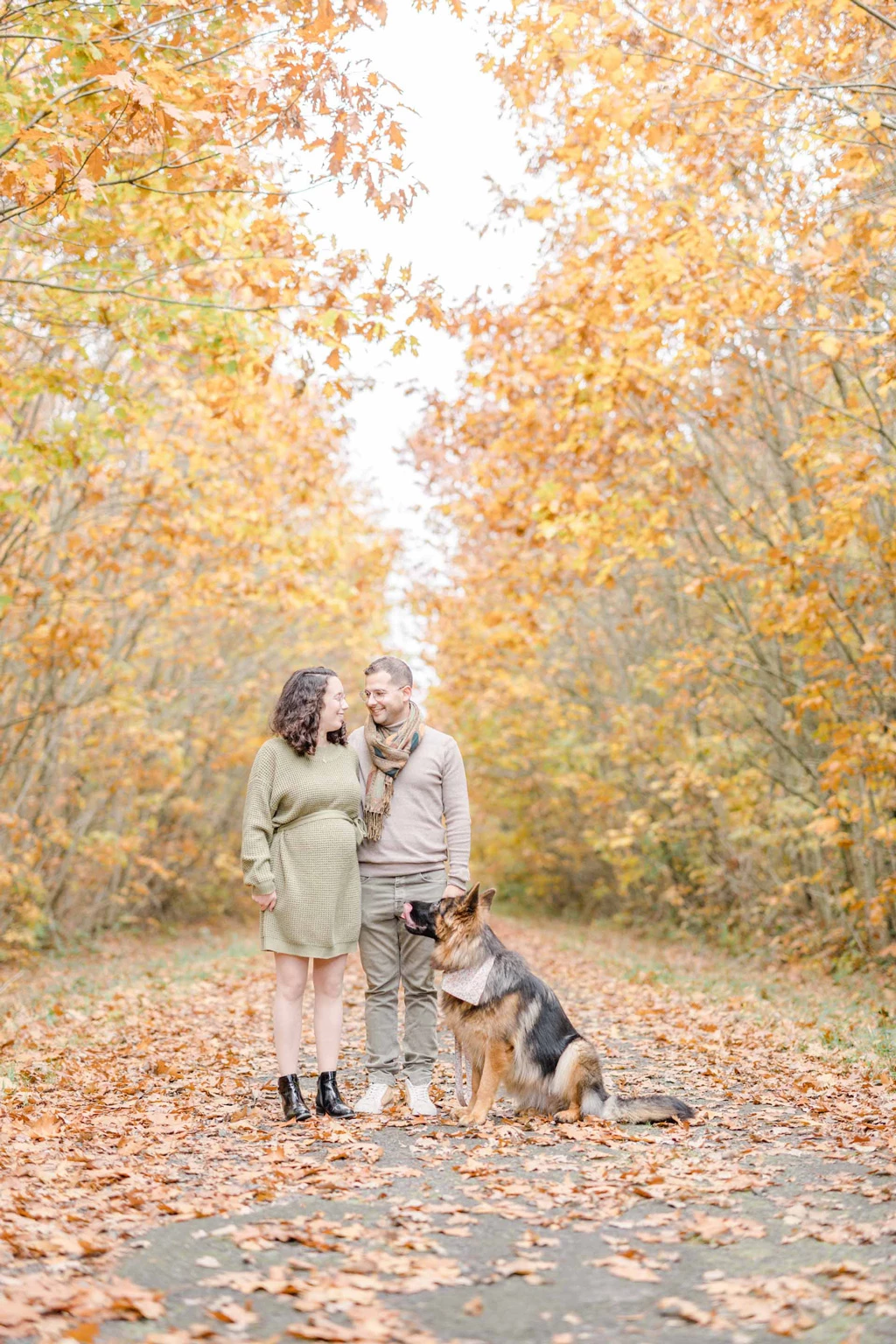 Séance photo famille automne : couple et chien en complicité sous une voûte de feuilles dorées, ambiance intemporelle et élégante