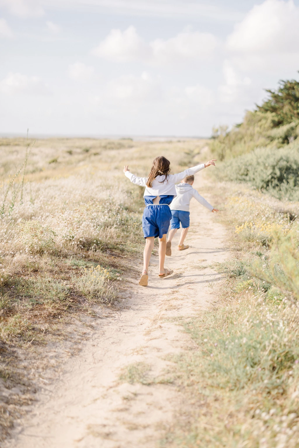 Deux enfants courant pieds nus sur un sentier de plage, bras écartés dans un élan de liberté joyeuse
