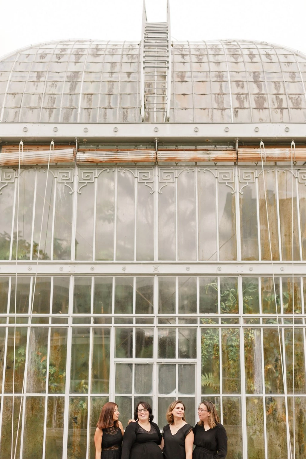 Séance photo de groupe élégante devant une serre historique : quatre femmes en tenues noires posent avec complicité et tendresse, baignées de lumière douce filtrée par les vitres.