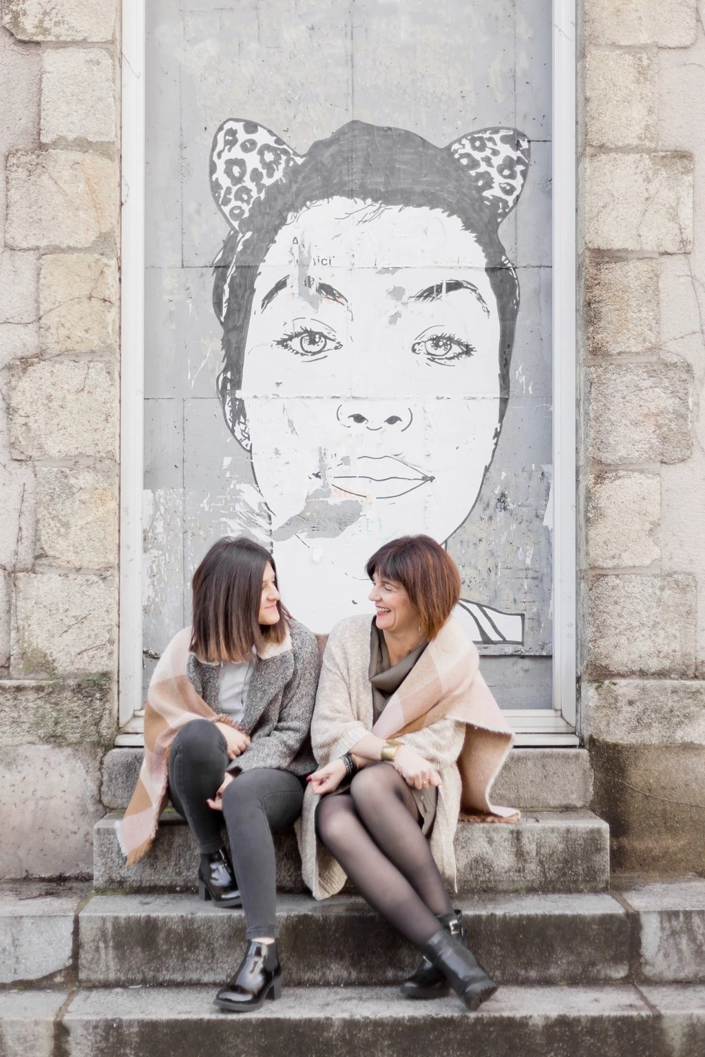 Deux femmes complices assises sur des marches en pierre devant une fresque murale urbaine