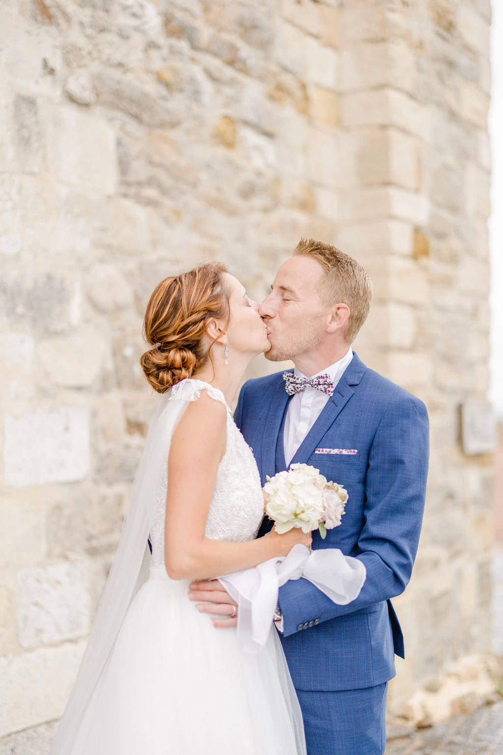 Couple de mariés s'embrassant devant un château en pierre, la mariée en robe blanche tient un bouquet