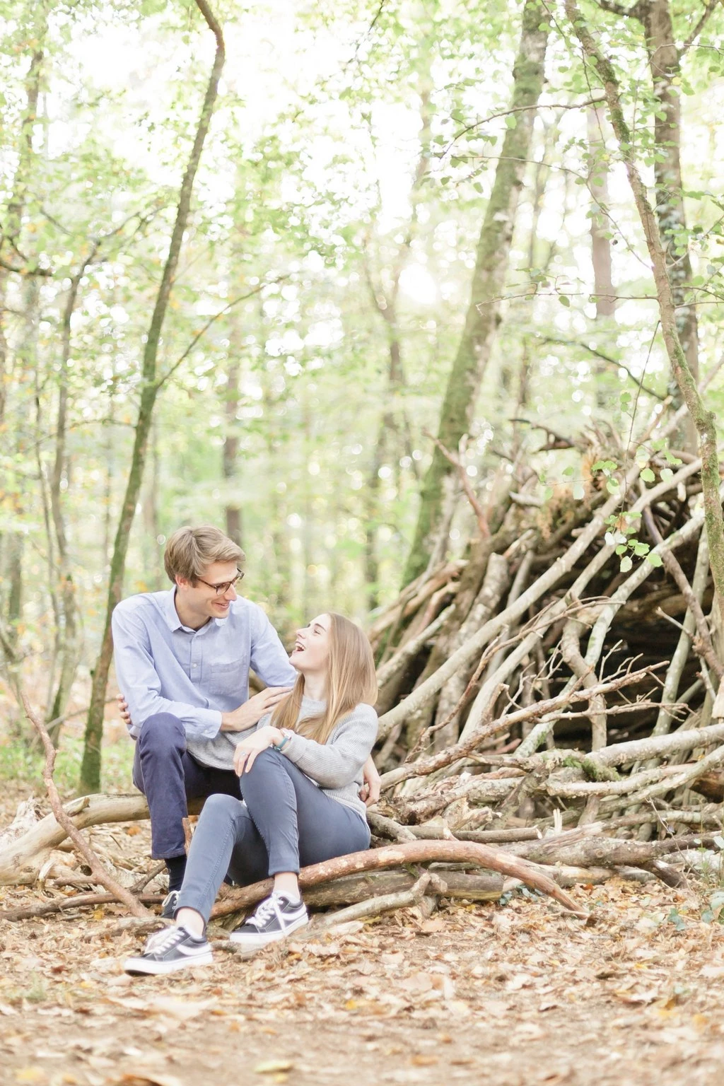 Séance couple en forêt : deux jeunes amoureux assis sur une branche, échangeant un regard tendre au milieu de la nature verdoyante, lumière douce et intemporelle
