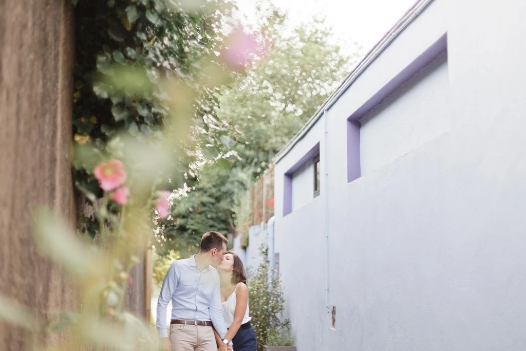 Séance famille en extérieur : couple marchant main dans la main dans une allée végétalisée, lumière dorée et fleurs roses en arrière-plan, ambiance tendre et intemporelle