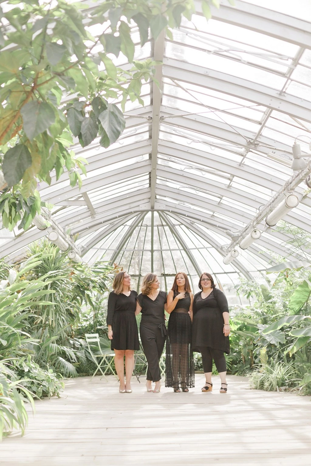 Séance photo famille en serre : quatre femmes en robes noires sous un parapluie transparent, complicité et tendresse dans la lumière douce d'une véranda végétale