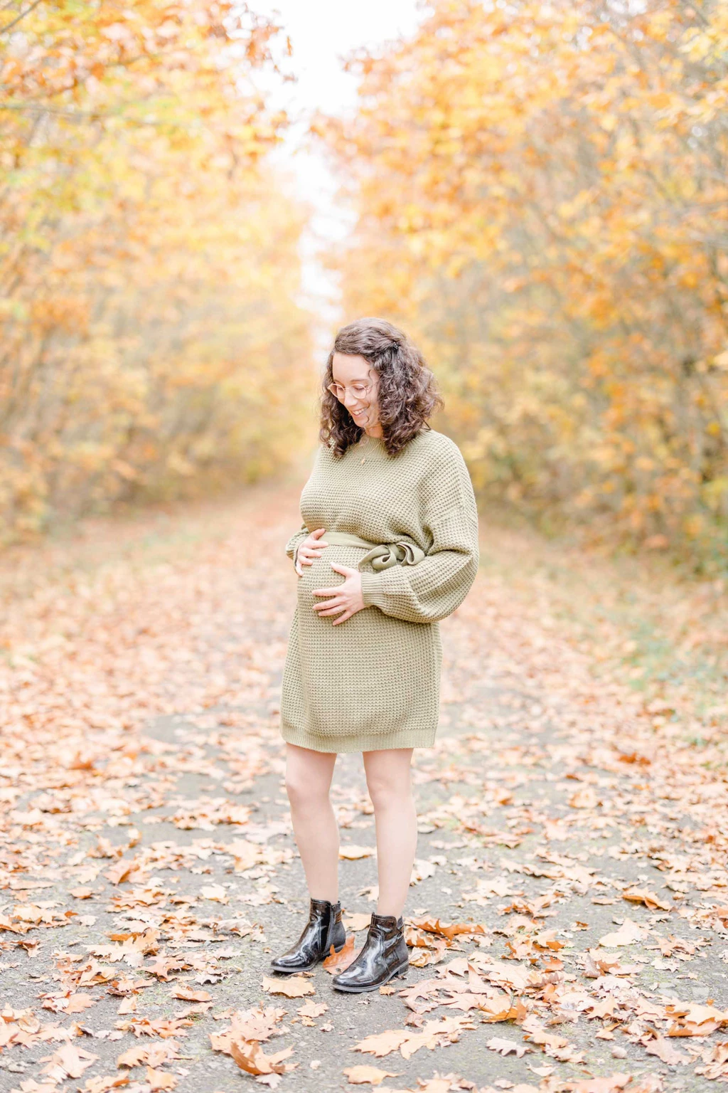 Future maman en robe beige caressant son ventre rond dans une allée forestière parsemée de feuilles d'automne