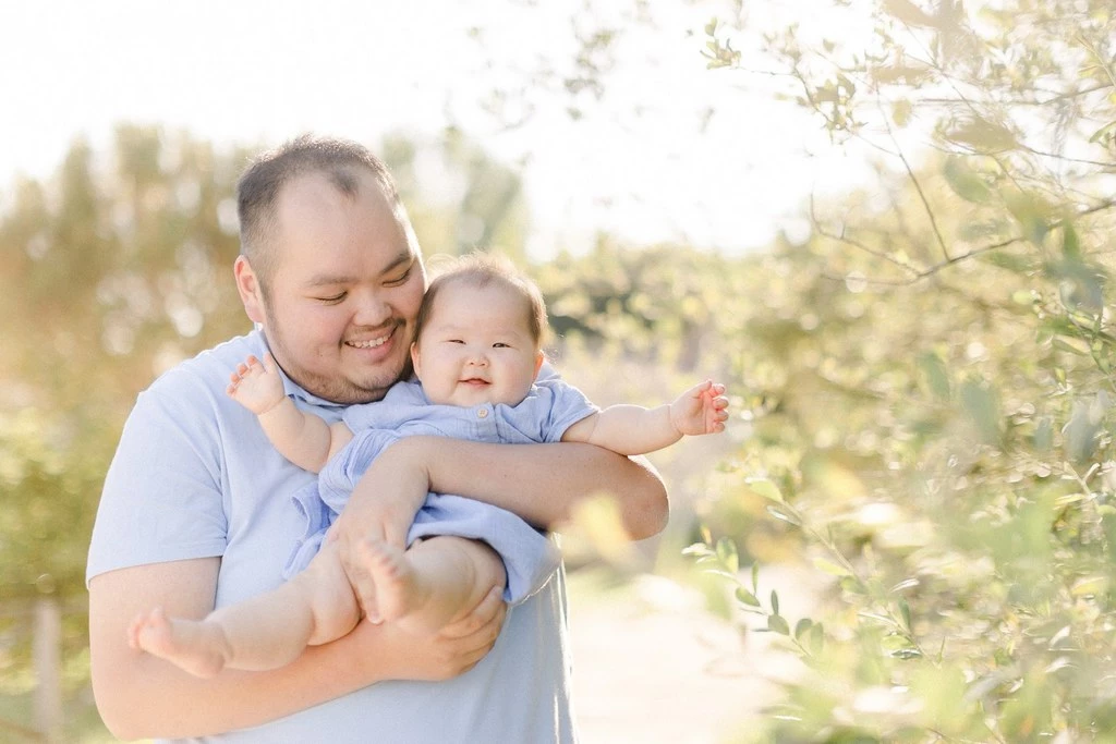 Séance photo famille en extérieur : père et bébé souriant dans la lumière dorée, tendresse et complicité intergénérationnelle, jardin fleuri au printemps