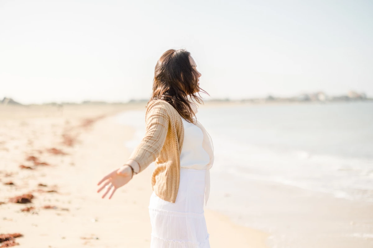 Femme  en robe blanche les bras ouverts sur une plage de sable au bord de l'eau
