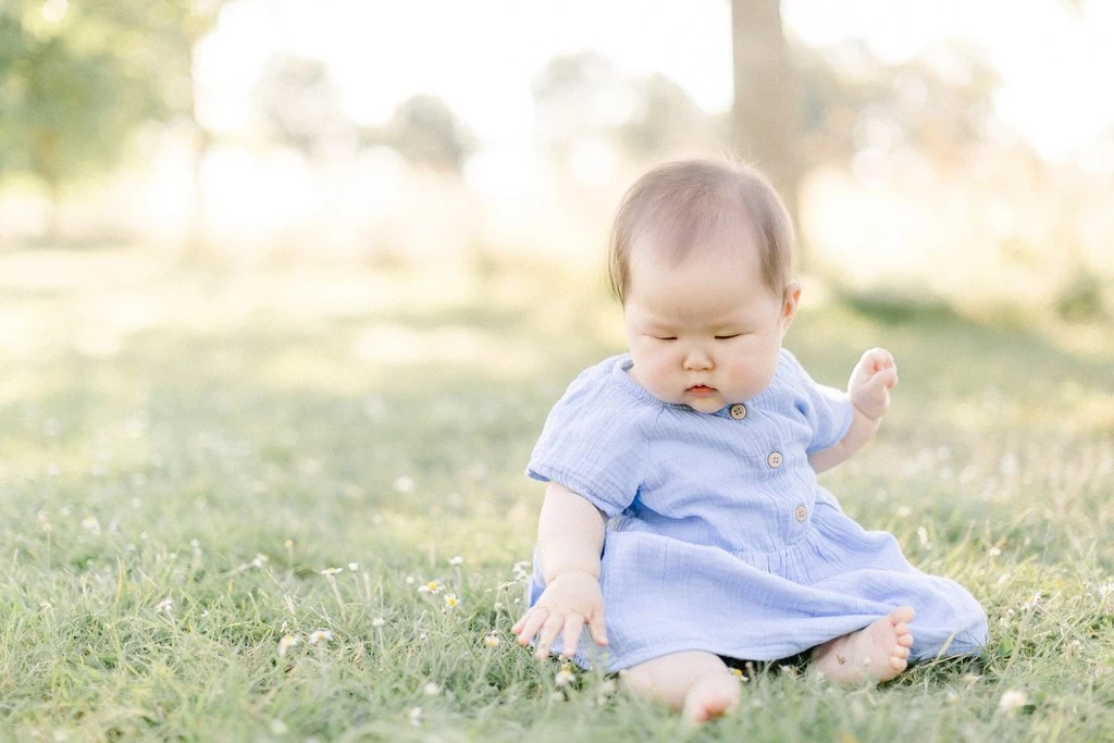 Portrait en extérieur d'un bébé en robe bleue assis dans l'herbe verte, lumière dorée naturelle, ambiance tendre et intemporelle