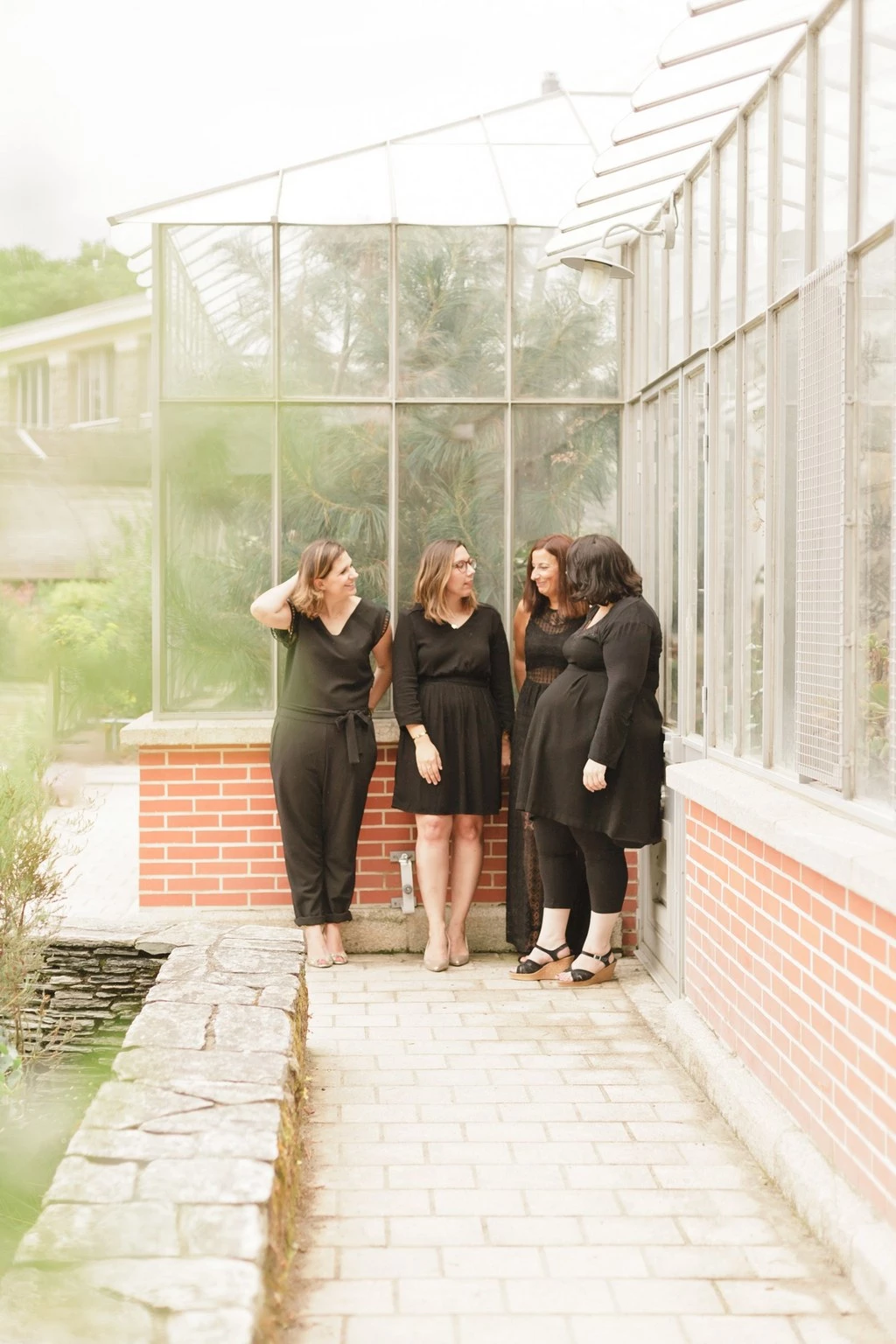 Trois femmes en tenue noire posent dans une véranda victorienne en brique rouge, baignées de lumière douce naturelle, exprimant une complicité tendre et authentique