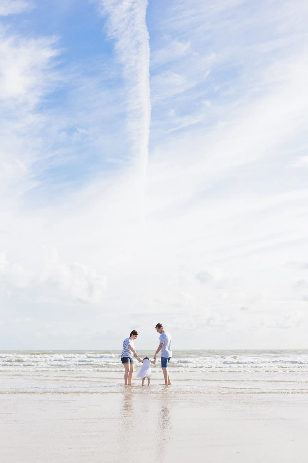 Séance famille à la plage : trois personnes en tenue claire se tenant la main dans l'eau, ambiance tendre et lumineuse en bord de mer vendéen