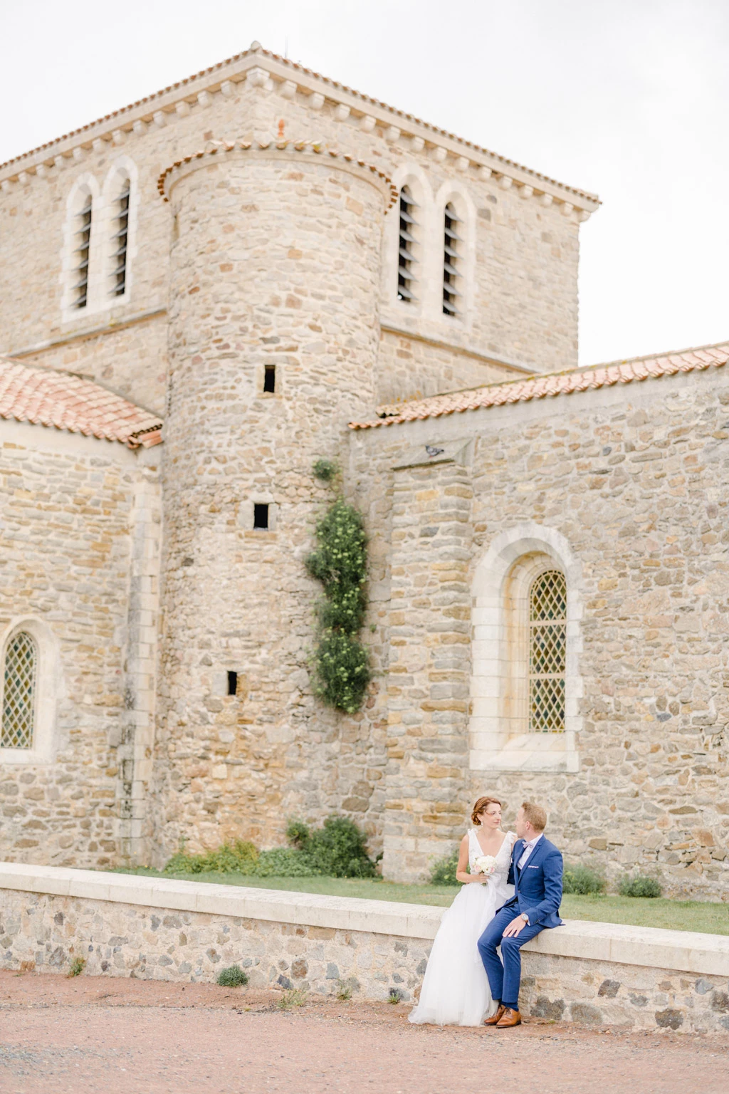 Couple de mariés assis devant une église romane en pierre, lumière douce et naturelle