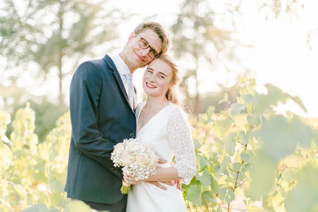 Couple de jeunes mariés enlacés dans une vigne ensoleillée, la mariée tenant un bouquet blanc