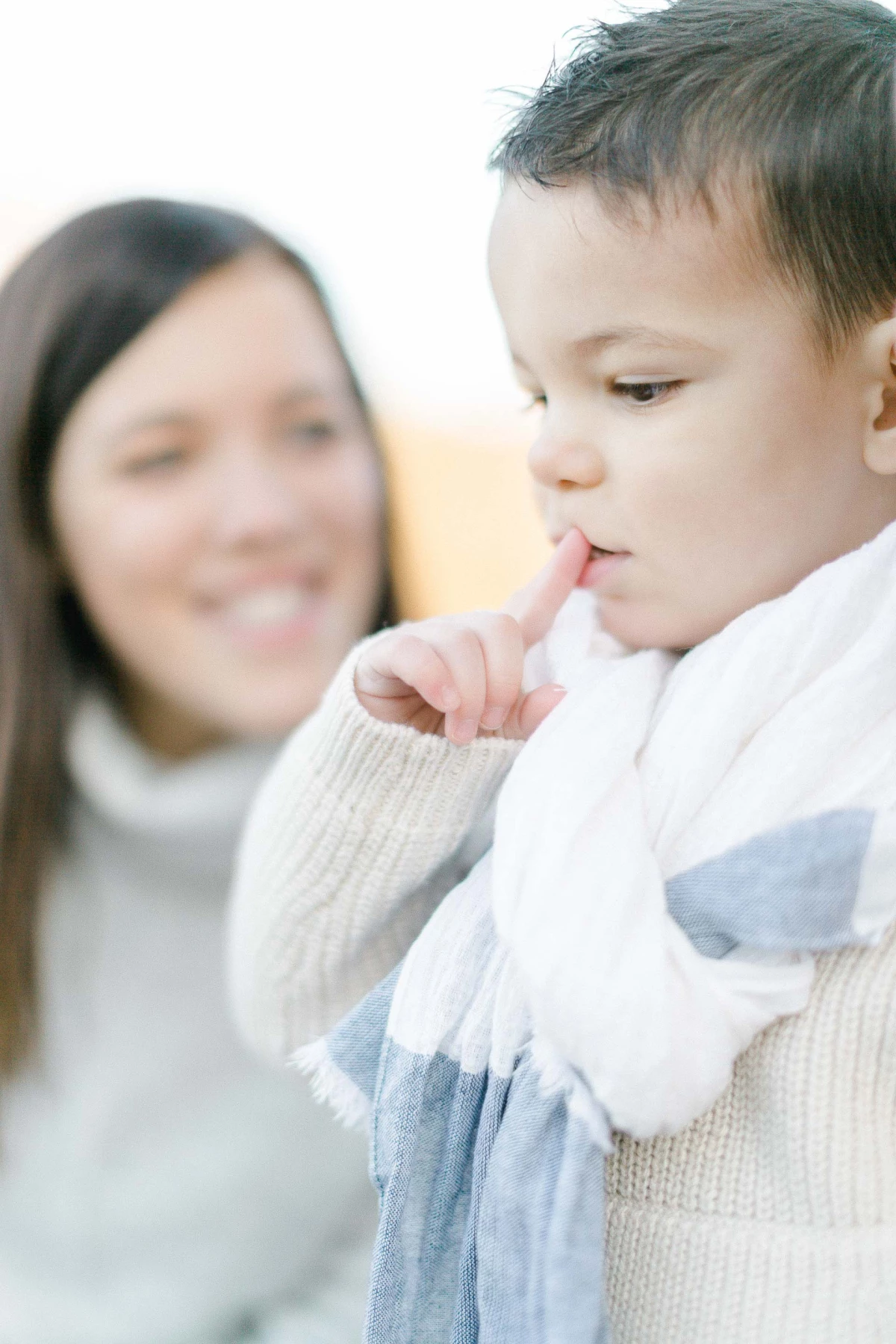 Séance photo famille : petit garçon en pull blanc avec femme floue en arrière-plan, tendresse et complicité dans une lumière douce et intemporelle