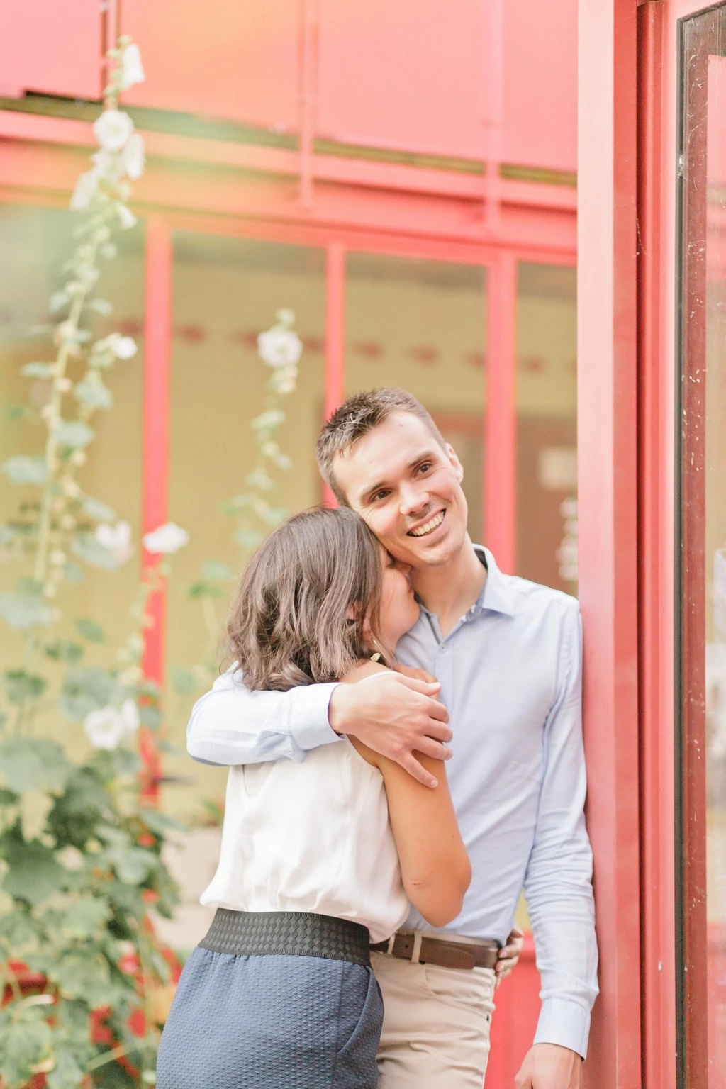 Séance portrait couple tendresse devant façade rose corail, homme en chemise bleue souriant embrassant femme en chemise blanche, ambiance douce et lumineuse