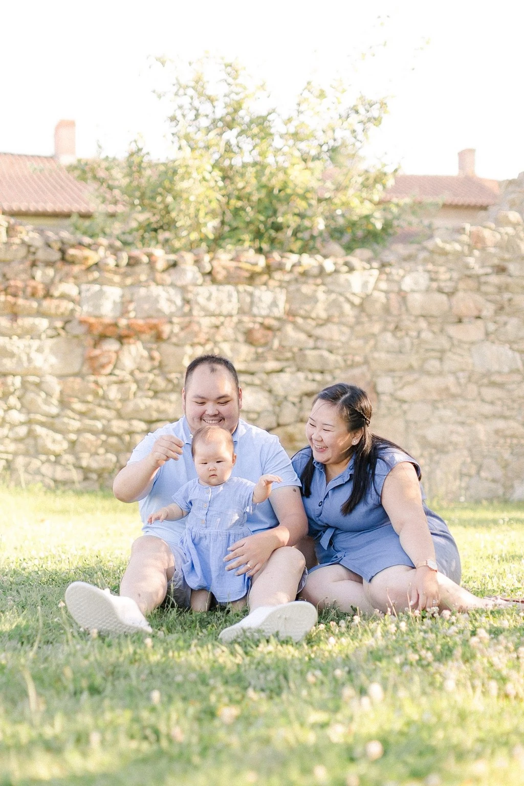 Séance photo famille en plein air : deux parents et leur enfant en bas âge assis dans l'herbe, vêtus de teintes bleues assorties, souriant avec tendresse devant un mur de pierre ancien