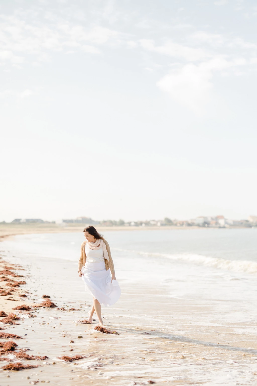 Mariée en robe blanche marchant pieds nus sur une plage au bord de l'eau, entourée d'algues échouées