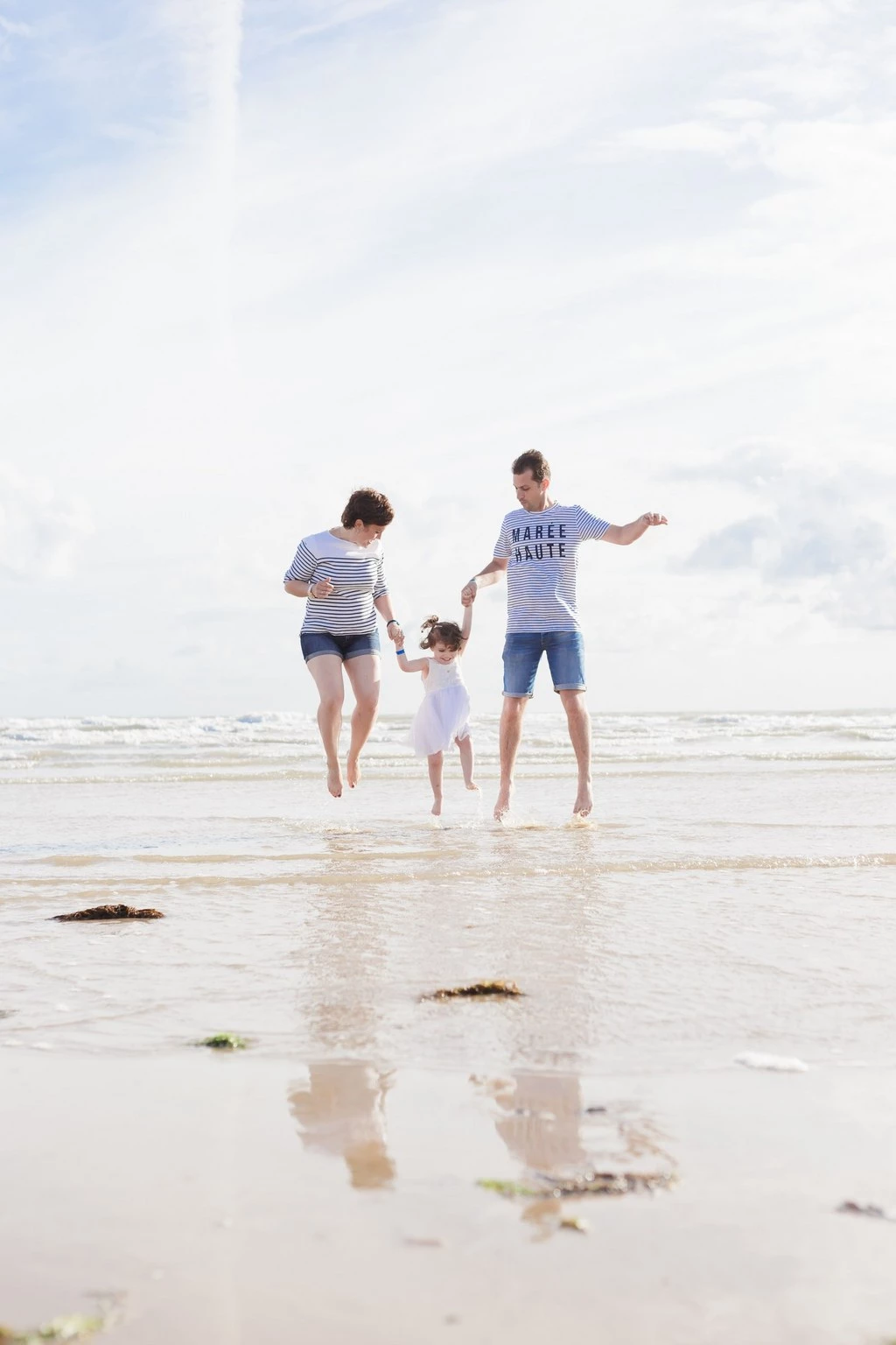 Séance photo famille à la plage : trois enfants courant pieds nus dans l'eau turquoise, complicité et joie, lumière douce et ciel nuageux en Vendée