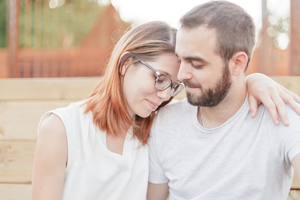 Séance couple tendresse : jeune femme aux cheveux roux et homme barbu en t-shirts blancs, enlacés dans une intimité complice, lumière douce dorée, ambiance intemporelle et raffinée