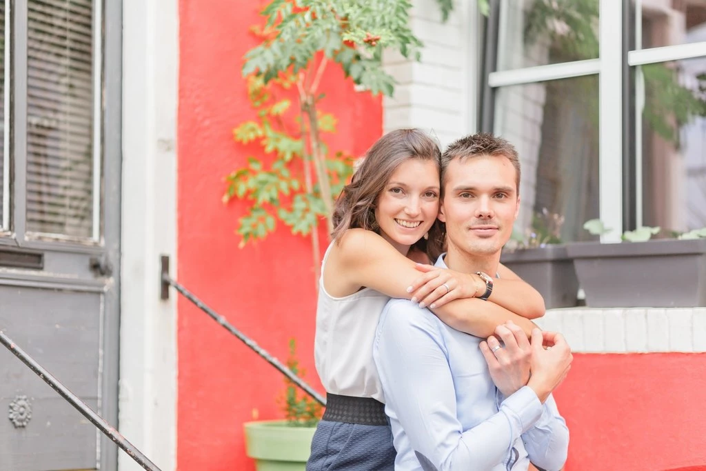 Séance couple tendresse : homme en chemise bleu ciel portant femme sur ses épaules, sourires complices devant façade rouge et blanche, lumière naturelle douce, intimité émotionnelle