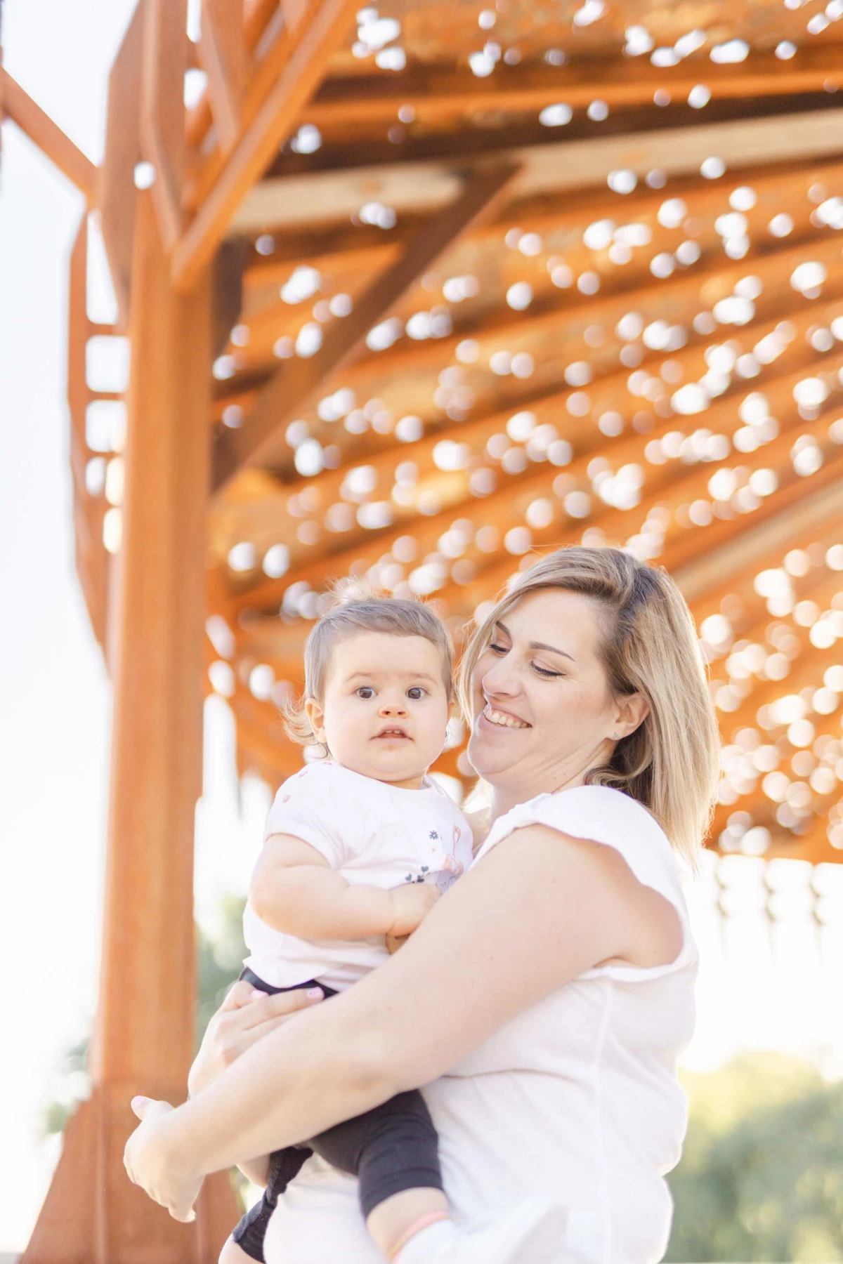 Séance photo famille : mère tenant son enfant en bas âge sous une pergola en bois avec guirlandes lumineuses, moment de tendresse et complicité