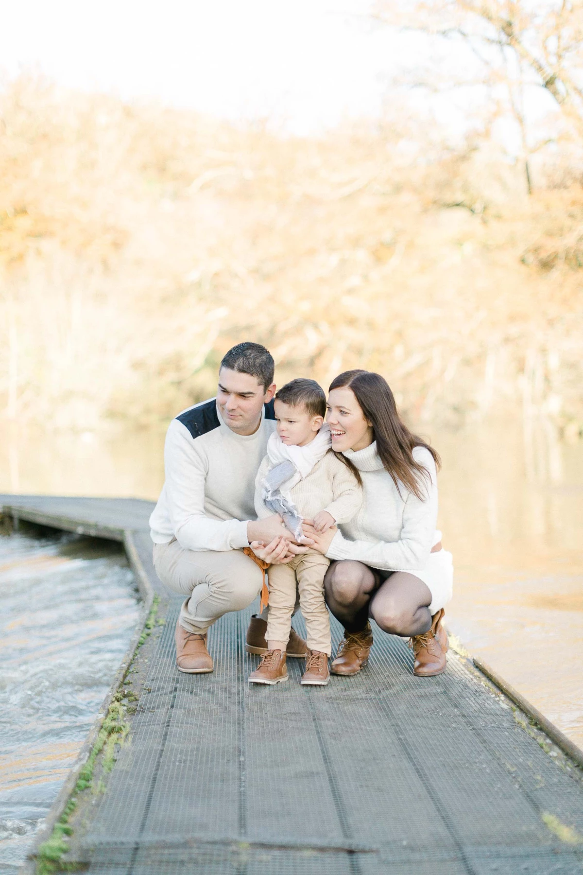 Séance photo famille complice sur une passerelle en bois : parents et enfant en tenue beige et blanche, lumière dorée d'automne, ambiance tendre et intemporelle en Vendée