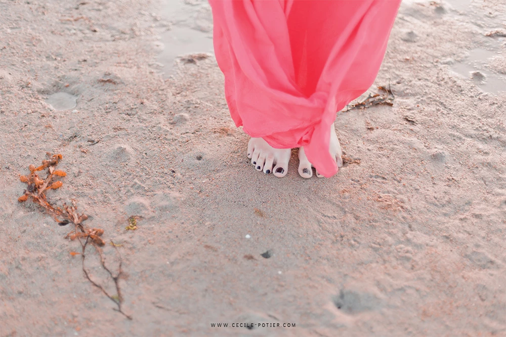 Séance photo maternité à la plage : détail des pieds nus d'une femme enceinte en robe rose poudré sur le sable, ambiance douce et intime