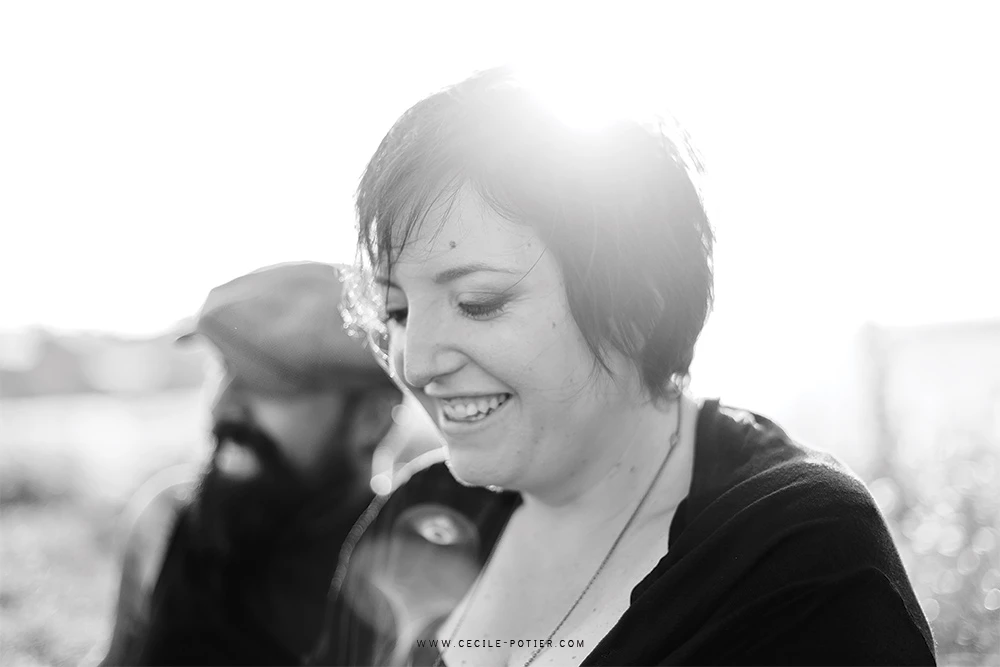 Portrait de couple en noir et blanc, femme souriante au premier plan, homme flou en arrière-plan, ambiance tendre et complice en extérieur, lumière douce et naturelle