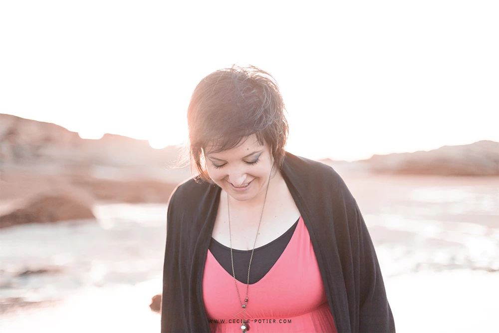 Portrait de femme en séance photo lifestyle en bord de mer, vêtue d'un top rose et cardigan noir, souriante dans la lumière dorée du coucher de soleil, ambiance émotionnelle et authentique