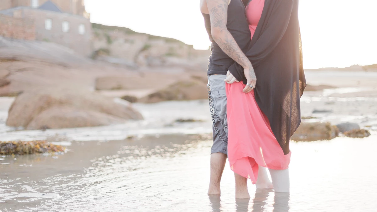 Séance photo famille à la plage : femme et enfant en robe rose marchant dans l'eau peu profonde, moment de tendresse et complicité au coucher de soleil