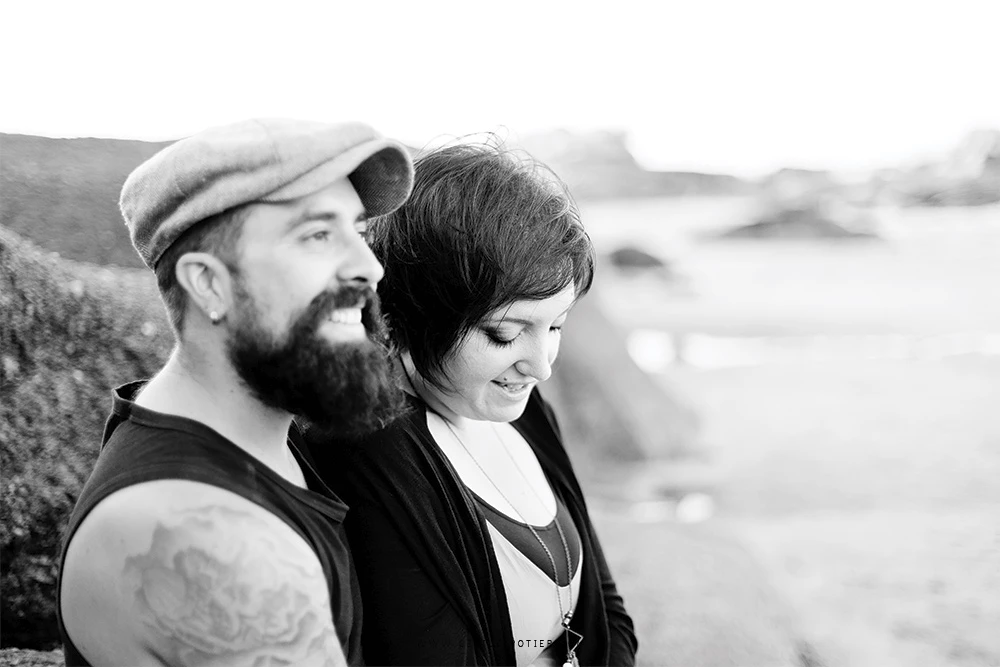 Portrait de couple en noir et blanc, homme barbu en casquette et femme souriante, complicité tendre en bord de mer, ambiance intemporelle et lumineuse