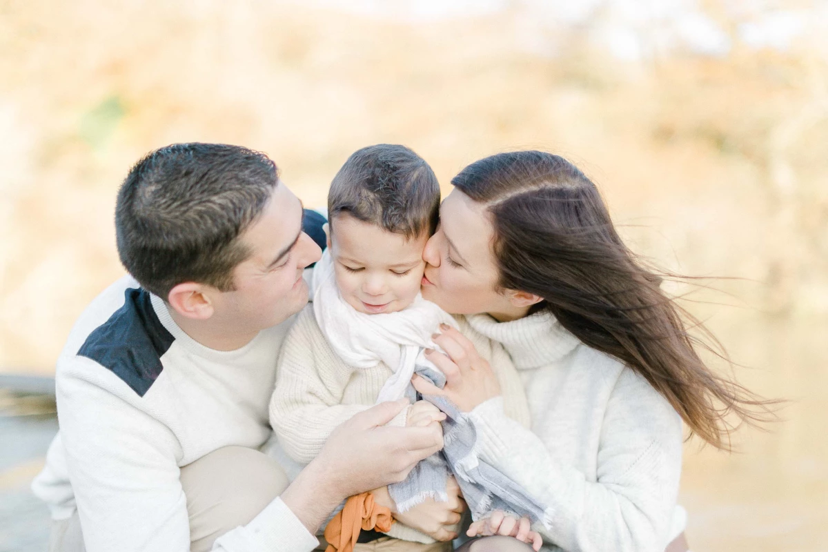 Séance famille portrait : parents embrassant tendrement leur jeune enfant, vêtus de blanc et beige, lumière dorée et douce, ambiance intemporelle et raffinée en extérieur