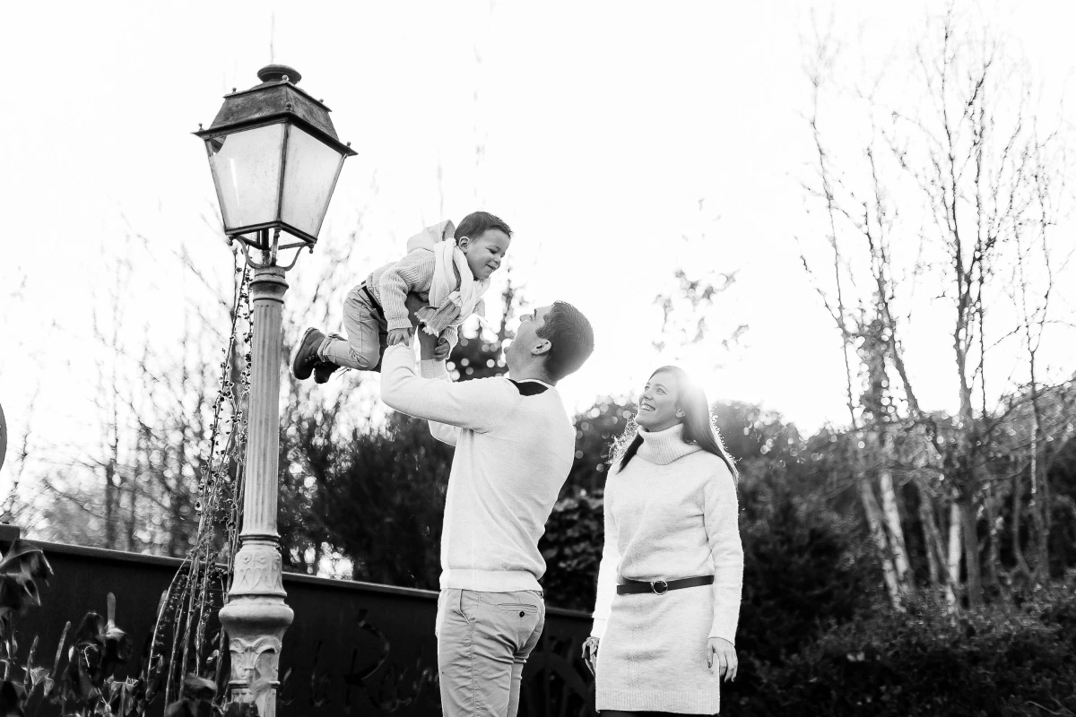 Séance photo famille en noir et blanc : père jouant avec son enfant sous un lampadaire vintage, femme souriante à côté, arbres nus en arrière-plan, ambiance tendre et intemporelle
