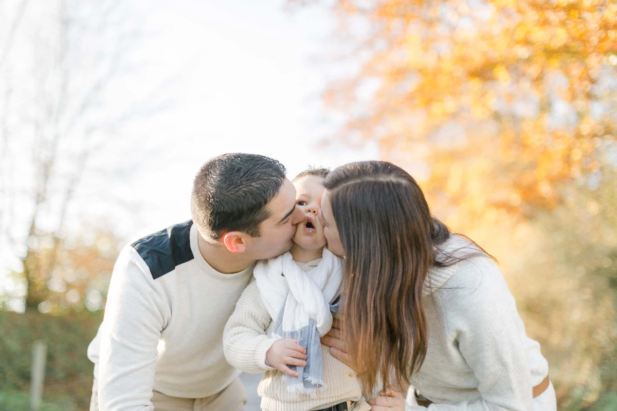 Séance photo famille en automne : parents embrassant tendrement leur enfant dans une lumière dorée, ambiance chaleureuse et intemporelle en pleine nature