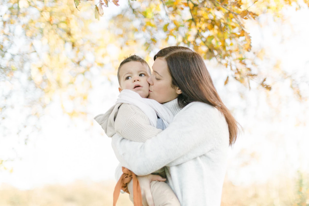 Séance photo famille en automne : mère embrassant tendrement son jeune enfant sous une pluie de feuilles dorées, lumière douce et chaleureuse
