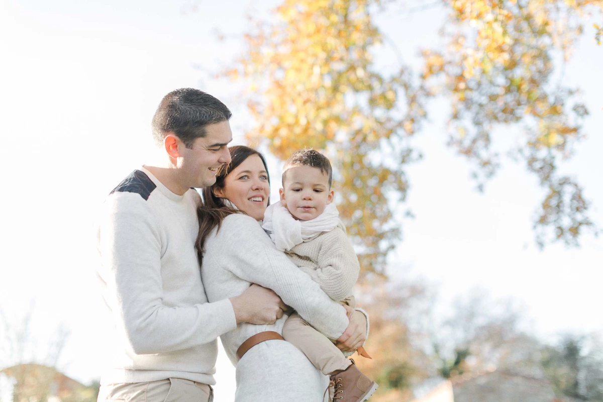 Séance photo famille tendresse : parents et enfant en pull beige souriant sous feuillage automnal doré, lumière douce et intimité authentique
