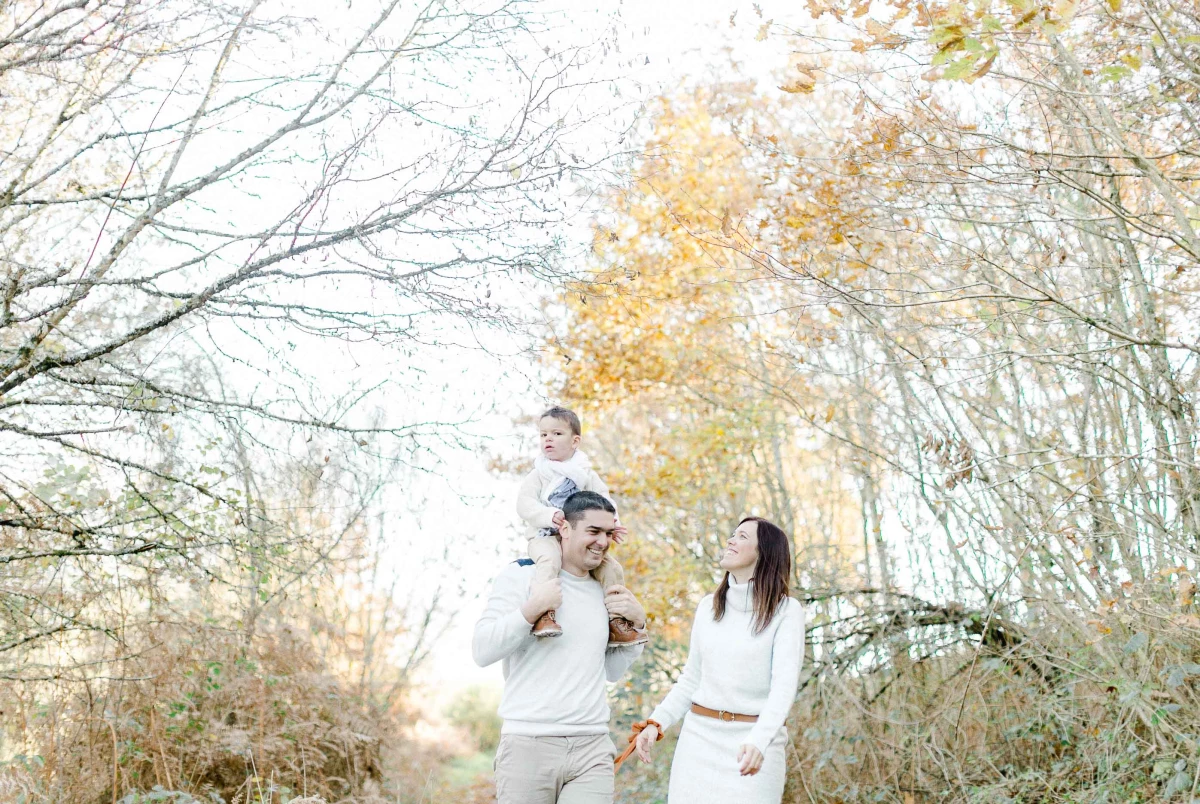 Séance photo famille en extérieur automnal avec père portant son enfant, mère souriante à ses côtés, ambiance chaleureuse et lumière dorée entre les arbres